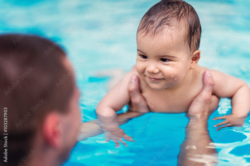 Ein Erwachsener hält ein lachendes Baby im Schwimmbad über Wasser. Das Baby hat nasse Haare und Wasserspritzer im Gesicht.