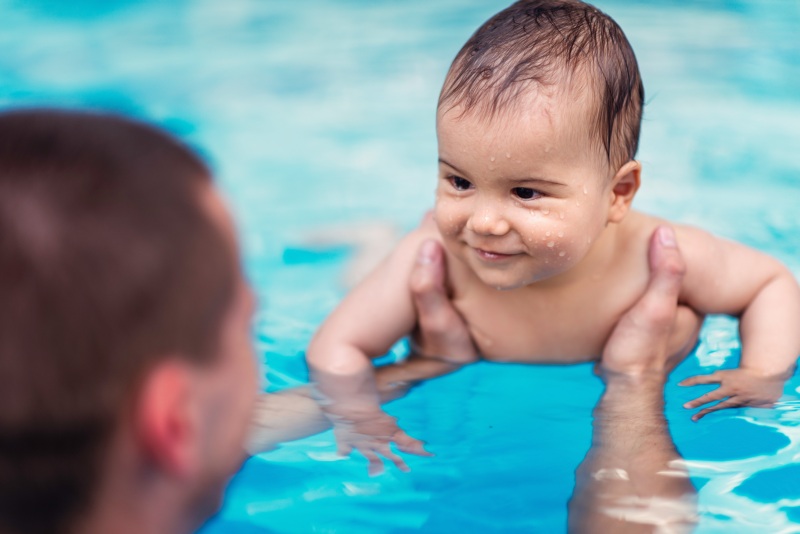 Babyschwimmen Ein Erwachsener hält ein lächelndes Baby im Wasser eines Schwimmbeckens. Das Baby ist nass und sieht glücklich aus.