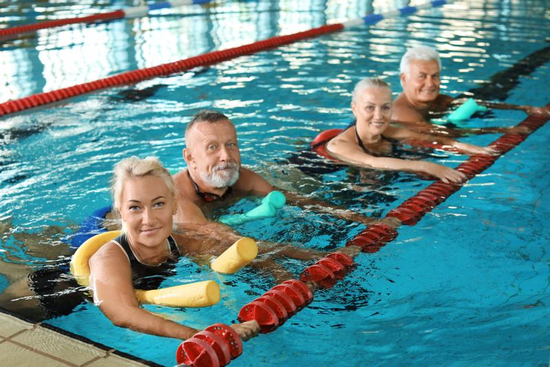 Wassergymnastik Vier ältere Erwachsene machen Wassergymnastik mit Poolnudeln in einem Schwimmbad und lächeln, während sie an der Beckenlinie festhalten.